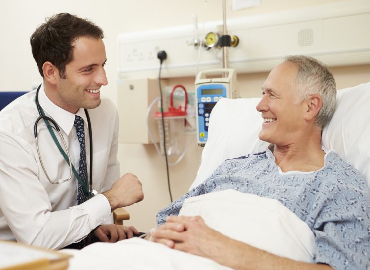 Doctor Sitting By Male Patient's Bed In Hospital